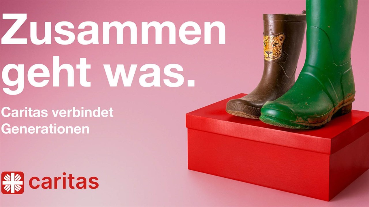 Auf einem roten Schuhkarton sind vor einem rosa Hintergrund ein Paar Gummistiefel zu sehen. Der linke Schuh ist ein brauner Kindergummistiefel mit einem Tigerkopf in kleiner Schuhgröße, der rechte ein hoher grüner Gummistiefel in großer Schuhgröße. Darüber steht das Kampagnenmotto: Zusammen geht was. Caritas verbindet Generationen.
