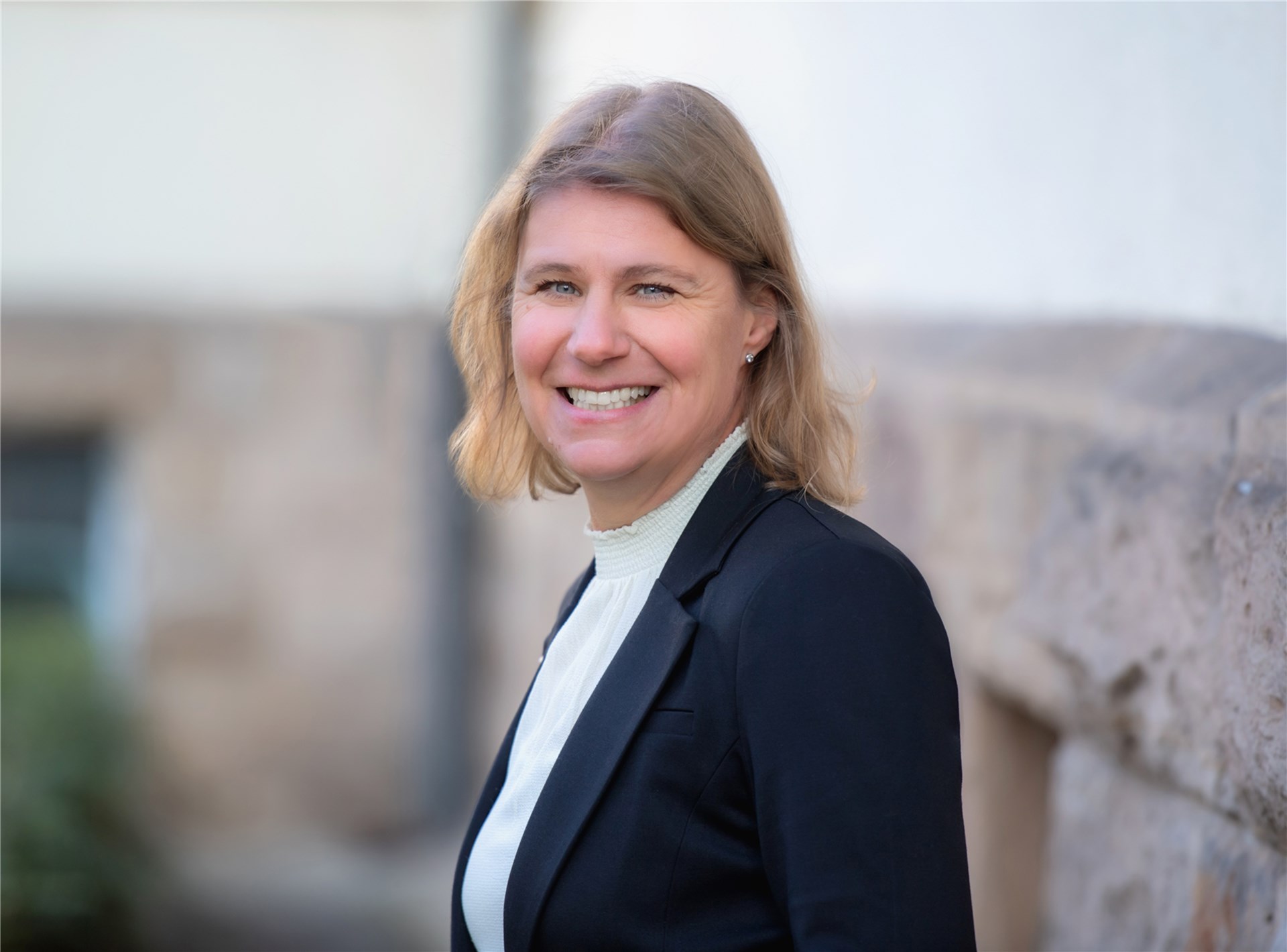 Eine Frau mit blonden Haaren steht im Zentrum des Bildes. Unter einem schwarzem Jacket trägt sie eine weiße Bluse mit Stehkragen. Der Hintergrund ist nicht erkennbar aber es ist irgendwo draußen. Sie lächelt den Betrachter an.