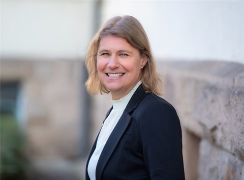 Eine Frau mit blonden Haaren steht im Zentrum des Bildes. Unter einem schwarzem Jacket trägt sie eine weiße Bluse mit Stehkragen. Der Hintergrund ist nicht erkennbar aber es ist irgendwo draußen. Sie lächelt den Betrachter an.