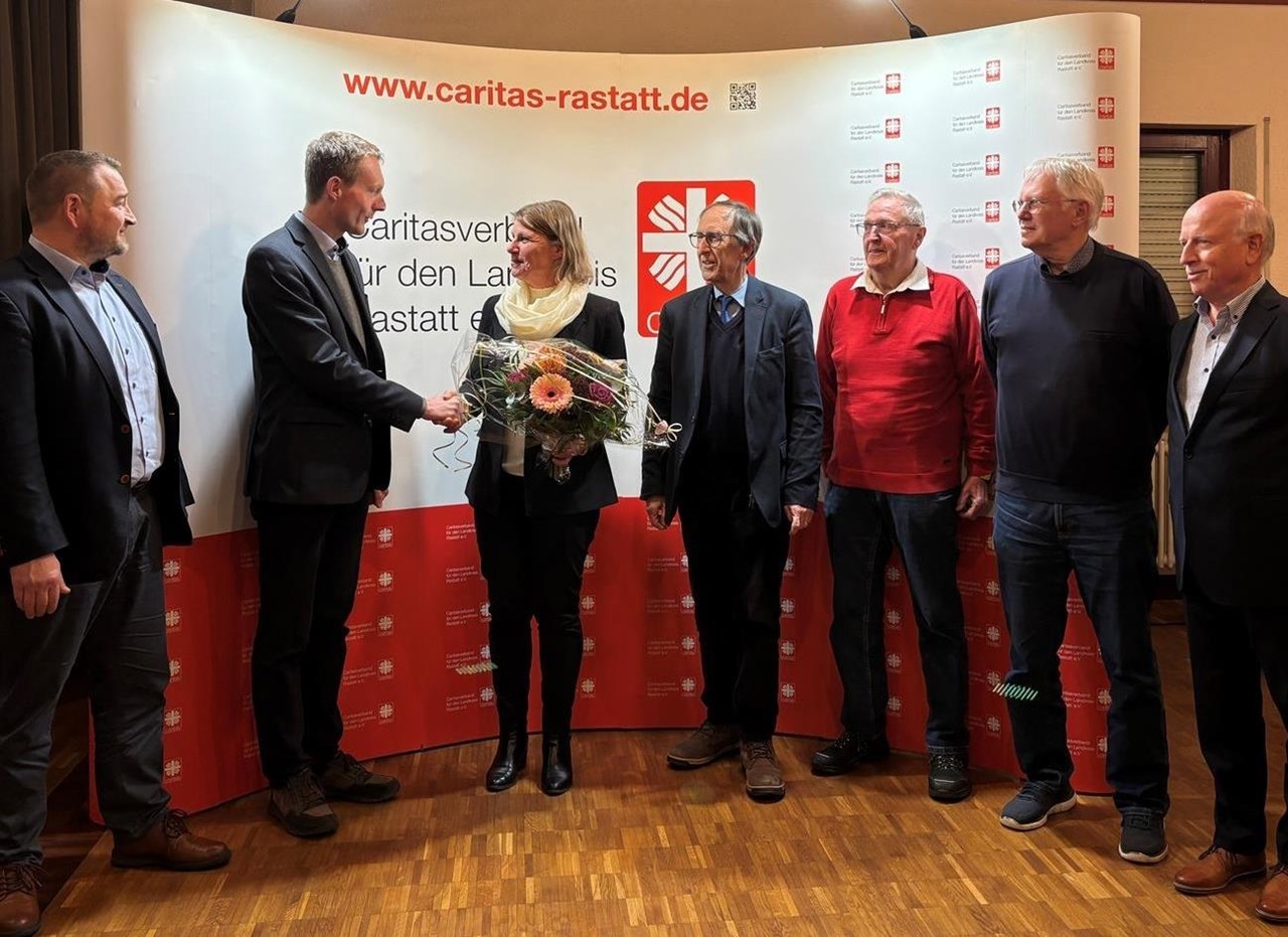Die Aufnahme zeigt eine Gruppe von sechs Personen, die in einem festlich vorbereiteten Raum vor einem Banner des Caritasverbandes für den Landkreis  Rastatt e.V. stehen. In der Mitte hält die neu gewählte Vorständin einen Blumenstrauß, während ihr von einer Person gratuliert wird. Die übrigen Personen stehen daneben und bilden eine kleine Halbkreisformation. Der Hintergrund ist geprägt von einem großen, weiß-roten Banner mit dem Verbands-Logo und der Website-Adresse „www.caritas-rastatt.de“. Der Raum hat eine warme Holzdecke und ist gut ausgeleuchtet, was eine feierliche und offizielle Atmosphäre vermittelt.