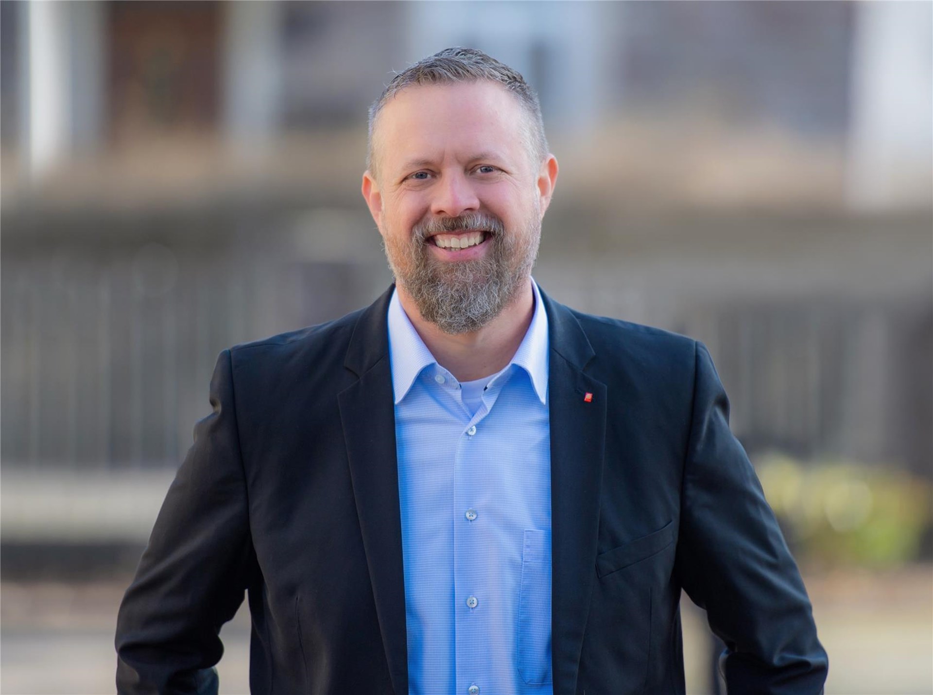 Ein Man mit bart schaur lächelnd den Betrachter an. Er hat ein helblaus Hemd an und eine dunkelblauen Sakko. Am Revers ist eine Caritasnadel zu sehen. Der Hintergrund ist nicht  erkennbar aber es ist irgendwo draußen.