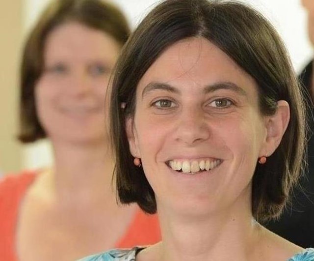 Das Foto zeigt eine Person im Vordergrund, die direkt in die Kamera blickt. Im Hintergrund sind drei weitere Personen zu sehen, die bewusst unscharf dargestellt sind. Die Aufnahme wirkt wie ein Porträt im Rahmen einer Team- oder Gruppenpräsentation. Durch die geringe Tiefenschärfe liegt der Fokus auf der Person im Vordergrund, während die weiteren Personen einen unterstützenden Hintergrund bilden.