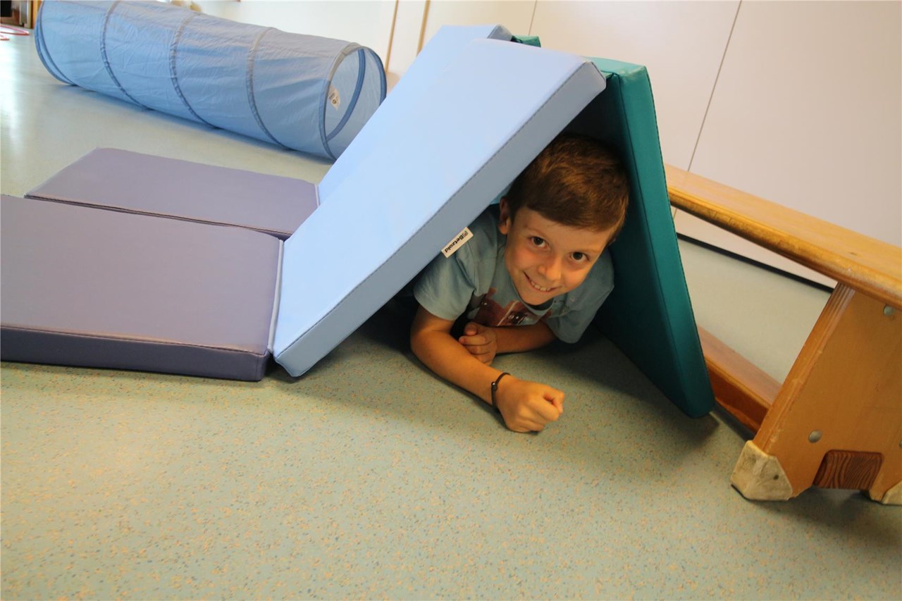 Ein Kind kriecht unter einer Höhle aus blauen Turnmatten hindurch, die kreativ zu einem kleinen Zelt aufgebaut wurden. Im Hintergrund ist ein blauer Spieltunnel zu sehen – alles auf einem hellblauen Boden in der Turnhalle des Kinderhauses Vimbuch.