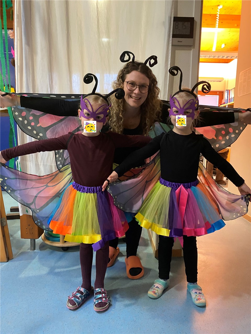 Vor einer jüngeren Frau mit Brille, die ein Schmetterlingskostüm trägt, stehen zwei kleine Mädchen im Kindergartenalter. Sie sind auch als Schmetterlinge verkleidet. Alle tragen masken und haben die Arme ausgebreitet.