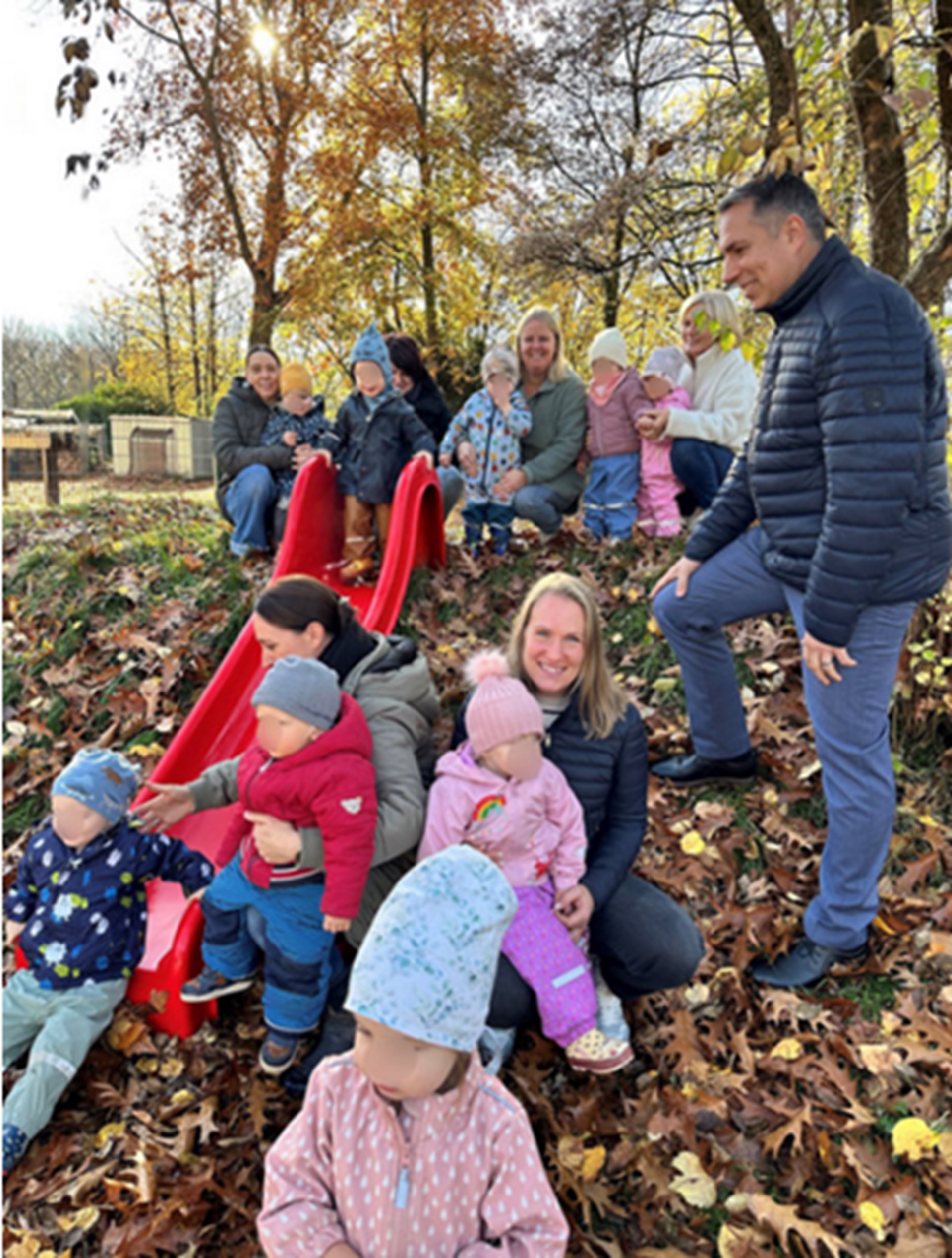 Das Foto zeigt eine Gruppe von Kleinkindern mit Erwachsenen im herbstlich bunten Außengelände eines Kindergartens. Im Mittelpunkt steht eine leuchtend rote Rutsche, die auf einem kleinen Hügel installiert ist und von vielen bunten Blättern umgeben ist. Einige Kinder sitzen am unteren Ende der Rutsche, andere stehen oder hocken daneben. Im Hintergrund sind weitere Kinder und Erwachsene zu sehen, die sich um die Rutsche gruppieren. Die Szene wirkt lebendig und fröhlich, mit warmem Sonnenlicht, das durch die Bäume fällt und die goldbraunen Herbstblätter zum Leuchten bringt. Die Kindergesichter wurden verpixelt.