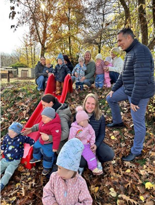 Das Foto zeigt eine Gruppe von Kleinkindern mit Erwachsenen im herbstlich bunten Außengelände eines Kindergartens. Im Mittelpunkt steht eine leuchtend rote Rutsche, die auf einem kleinen Hügel installiert ist und von vielen bunten Blättern umgeben ist. Einige Kinder sitzen am unteren Ende der Rutsche, andere stehen oder hocken daneben. Im Hintergrund sind weitere Kinder und Erwachsene zu sehen, die sich um die Rutsche gruppieren. Die Szene wirkt lebendig und fröhlich, mit warmem Sonnenlicht, das durch die Bäume fällt und die goldbraunen Herbstblätter zum Leuchten bringt. Die Kindergesichter wurden verpixelt.