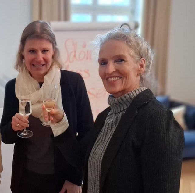 Zwei Frauen sehen in die Kamera. Die im Vordergrund hält ein Sektgals in der Hand und es sieht so aus, als wolle sie dem betrachter zuprosten. Auch die frau im Hintergrund hält ein Glas.