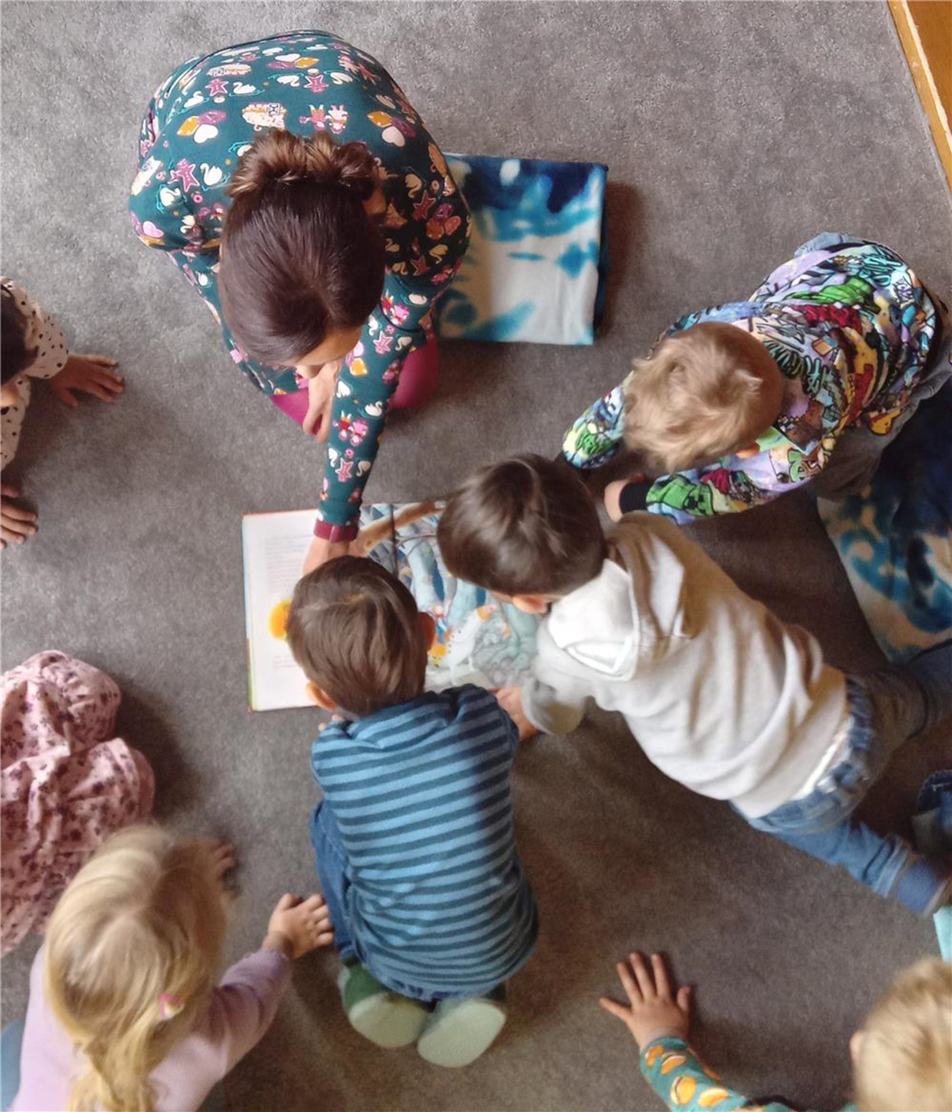 Das Foto zeigt eine Gruppe von Kindern und eine Erwachsene von oben im Kreis sitzen die sich über ein aufgeschlagenes Buch beugen.