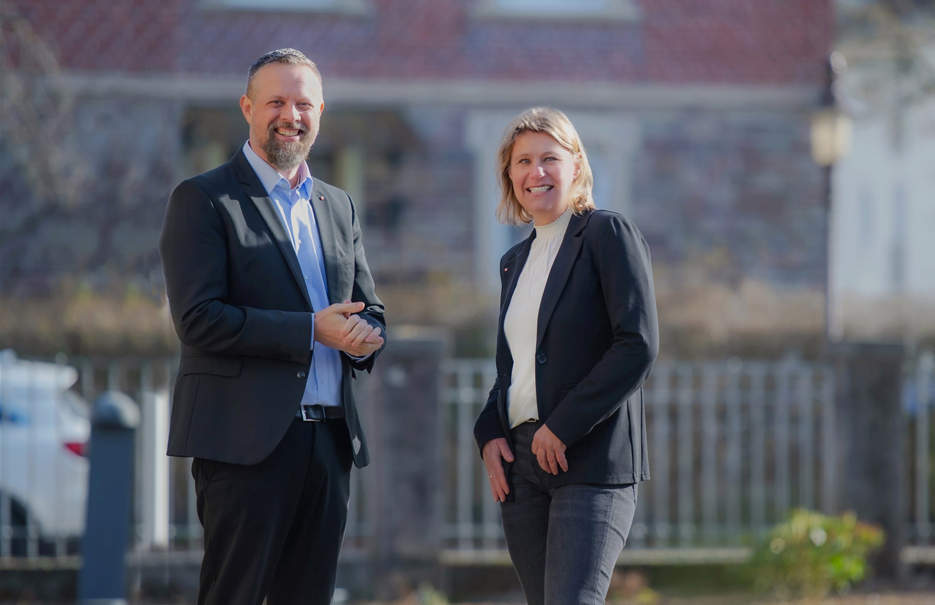 Ein Mann mit Bart und eine blonde Frau stehen auf einem freien Platz und lachen den betrachter an. Im Hintergrund ist ein Zaun schemenhaft zu shen. beide tragen dunkle Sakkos. Beide haben eine Caritasnadel im rever stecken. Der Mann hat ein blaus hemd an und hlät die Hände zusammen for seinem Körper. Die Frau hat die Arme locker an den Seiten. 